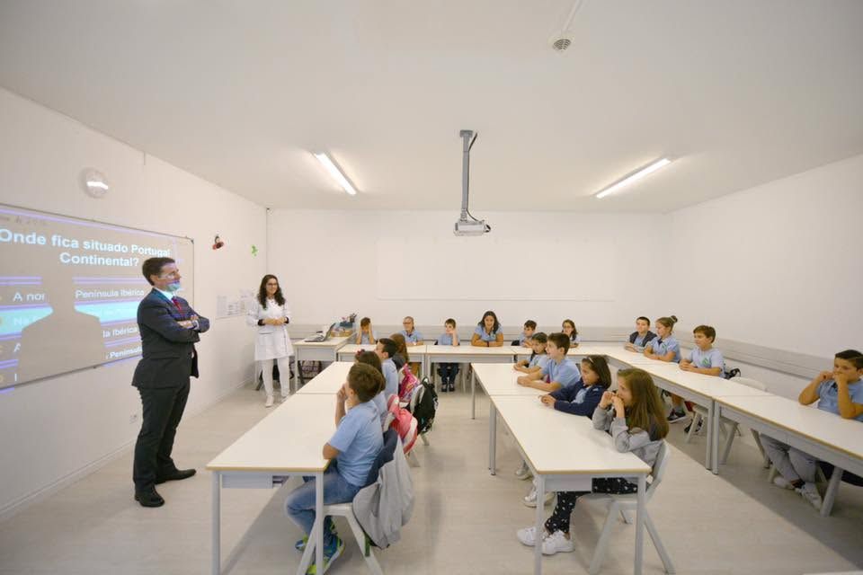 A large, bright classroom with pupils seated at their desks and teachers by the interactive whiteboard