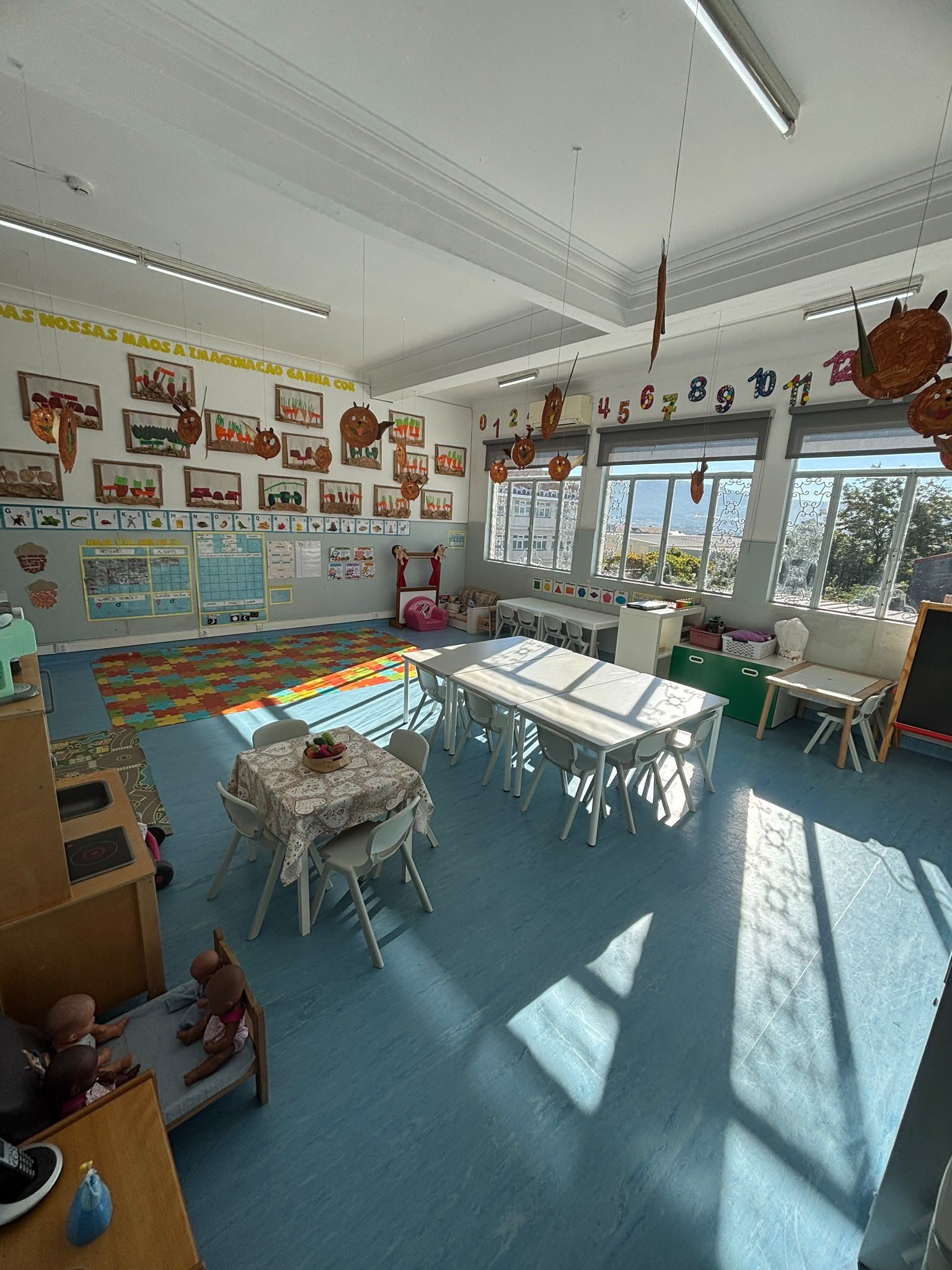 Sala de atividades do Pré-escolar, com mesas pequenas, cantinho da casinha e trabalhos das crianças nas paredes