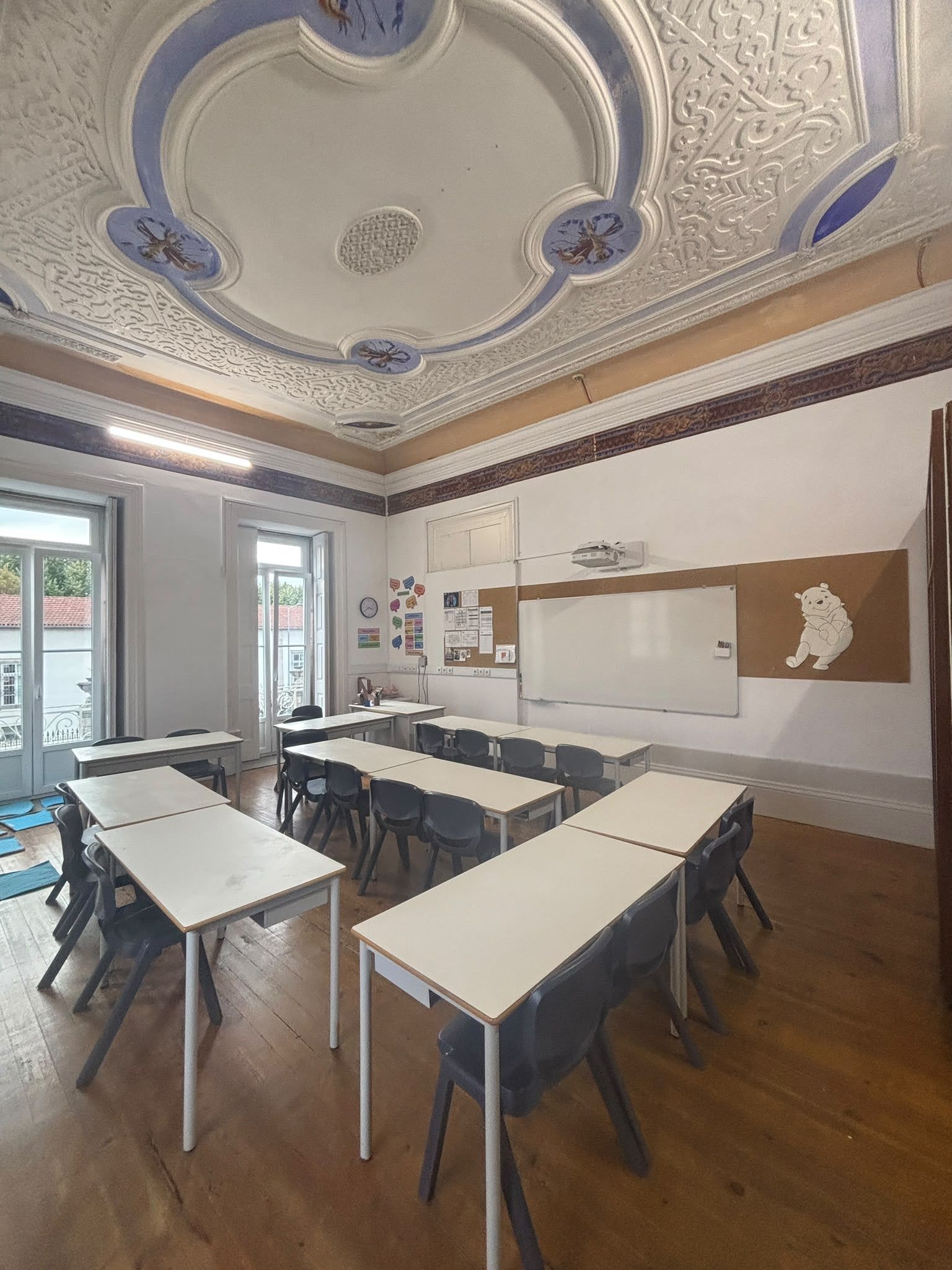 A classroom with an ornate old plaster ceiling, white desks and tall windows